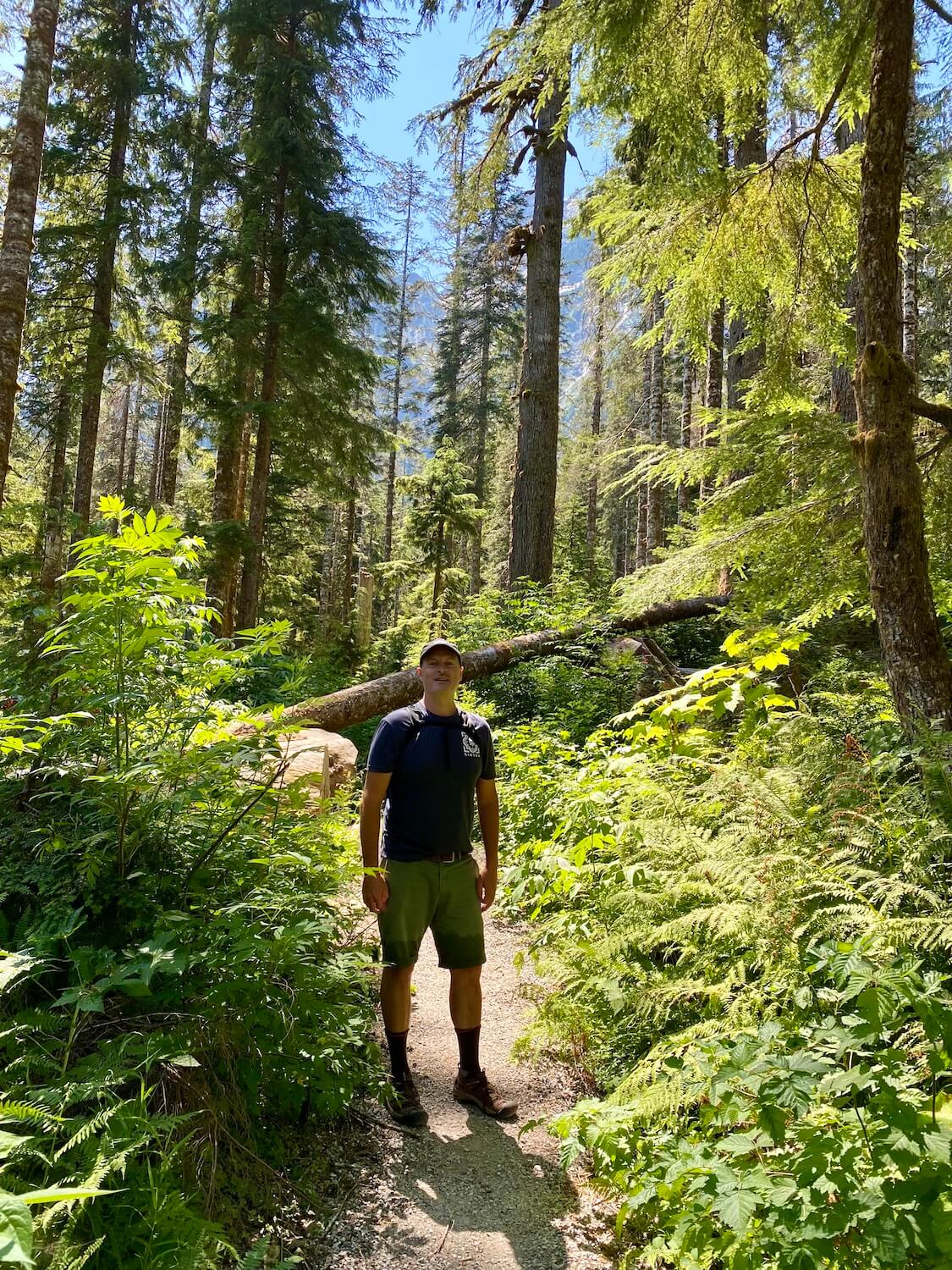 Matthew Kessi poses for a photo in the middle of a rich green forest on the trail to the Big Four Ice Caves in the Mt Baker Snoqualmie National Forest. He's wearing green shorts and a blue t-shirt and a black cap and there are many varieties of shrubs and trees around him with patches of blue sky peeking from above.