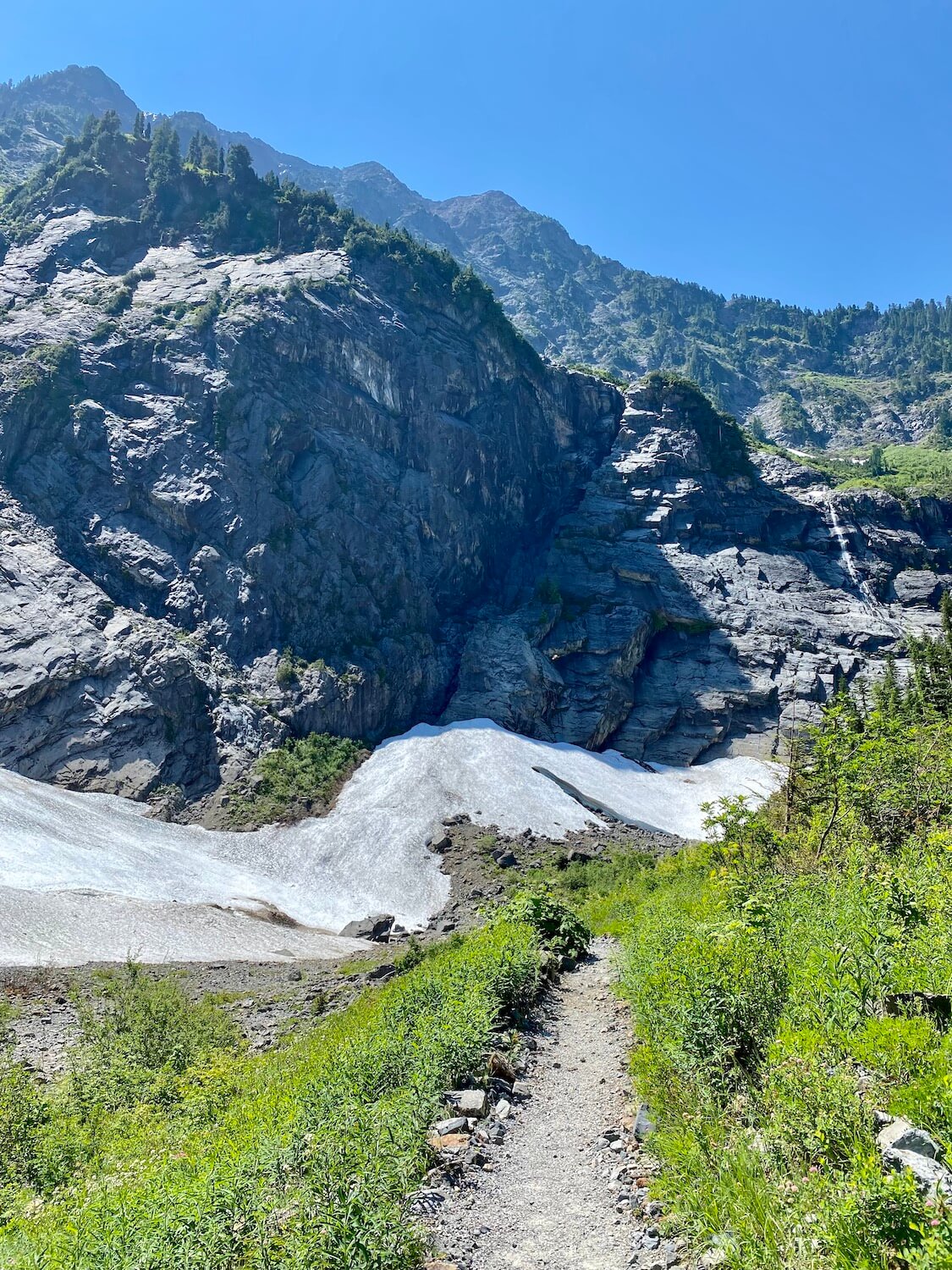 Big Four Mountain takes a commanding position in this photo of a gravel hiking trail leading toward a field of snow and ice at the base of the granite mountain.