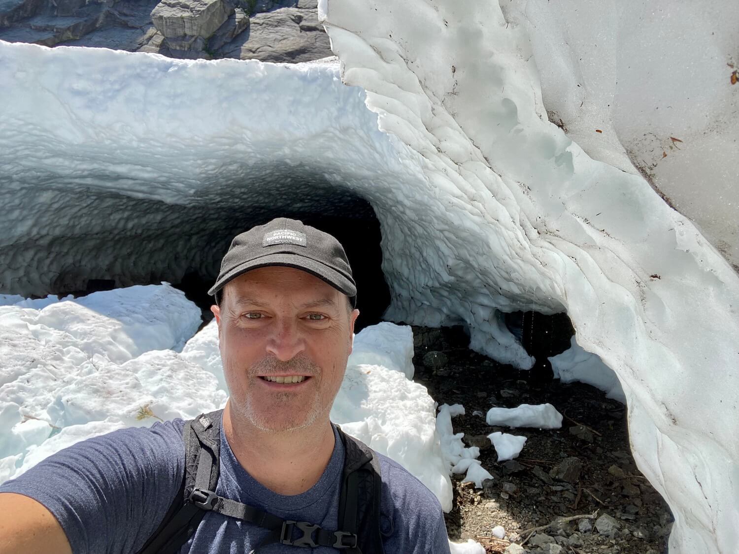 Matthew Kessi poses for this selfie in front of one of the caves at Big Four Ice Caves in the Mt. Baker Snoqualmie Wilderness Area. He's wearing a black cap and blue t-shirt and smiling toward the camera. The cave formations behind him are made from ice that is white with traces of dust and dirt from the rock below.