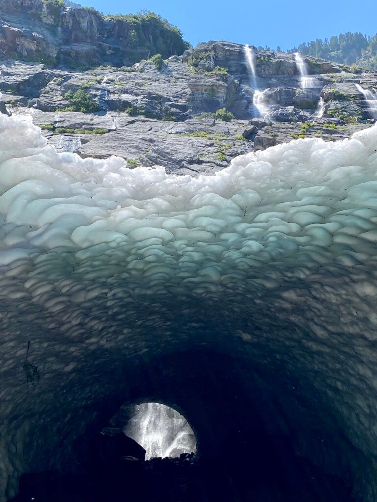 One of the ice caves at Big Four Ice Caves. In this shot a waterfall can be seen deep inside the cave, that appears dark leading out to the beveled roof of ice that shows blueish tints toward the opening of the cave. High above are mountain waterfalls flowing down the face of the sheer cliffs.