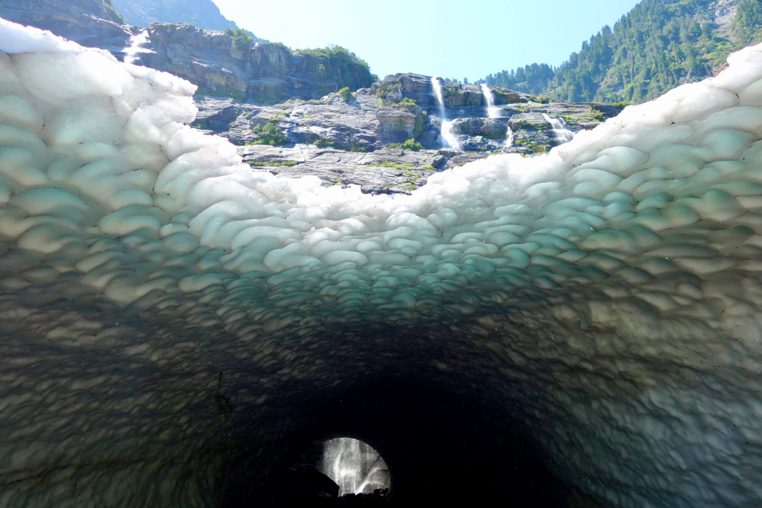 One of the impressive ice caves at Big Four Ice Caves showcases the towering mountains with waterfalls cascading down the chucks of rock amongst green shrubs. The ice cave has a waterfall in the background and the ceiling of the cave has many divers from the melting ice.