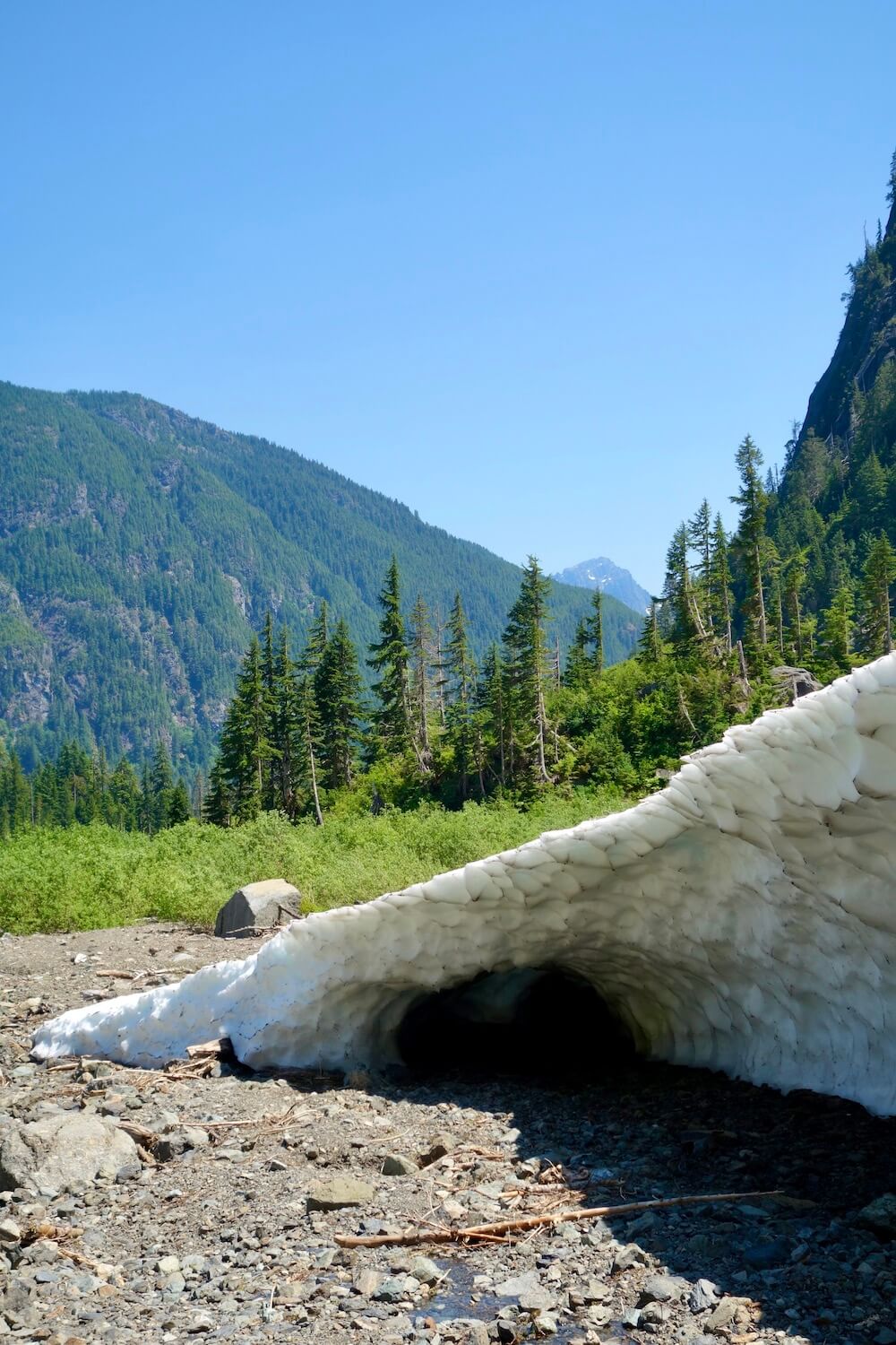 A view looking out from one of the caves made from ice and snow at the Big Four Ice Caves. The snow makes a diagonal line toward the rocks at the valley floor while fir trees pop up in the forest in the distance. The hills farther away roll with a rocky peak popping up in the background.