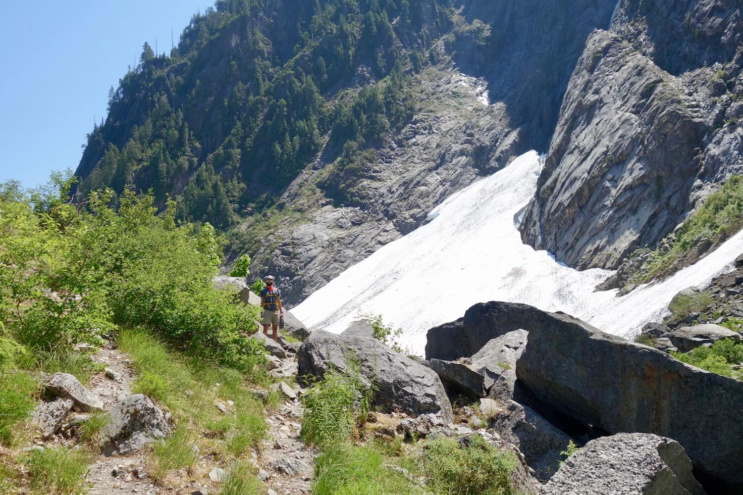 A hiker works through a rock scramble with huge jagged pieces of granite on the valley floor near the snow and ice field and the steep towering mountain.