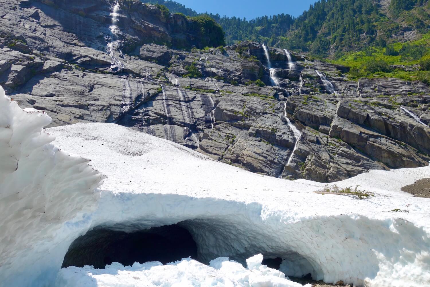 Big Four Ice Caves view looking up the mountain with alpine waterfalls flowing down the face of the mountain surrounded by green grass and foliage. Meanwhile the cave at the valley floor is made from snow and ice.