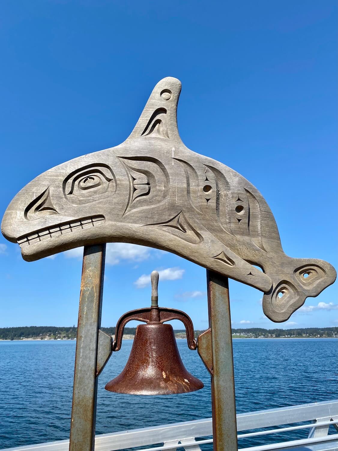 A native carving depicting an orca whale proudly displays on top of a copper colored bell on a pier in Coupeville, Washington on Whidbey Island.