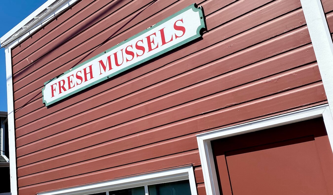 A large white sign with red lettering reads "Fresh Mussels" and is affixed to a painted red panels on the side of a building near the wharf in Coupeville, Washington.