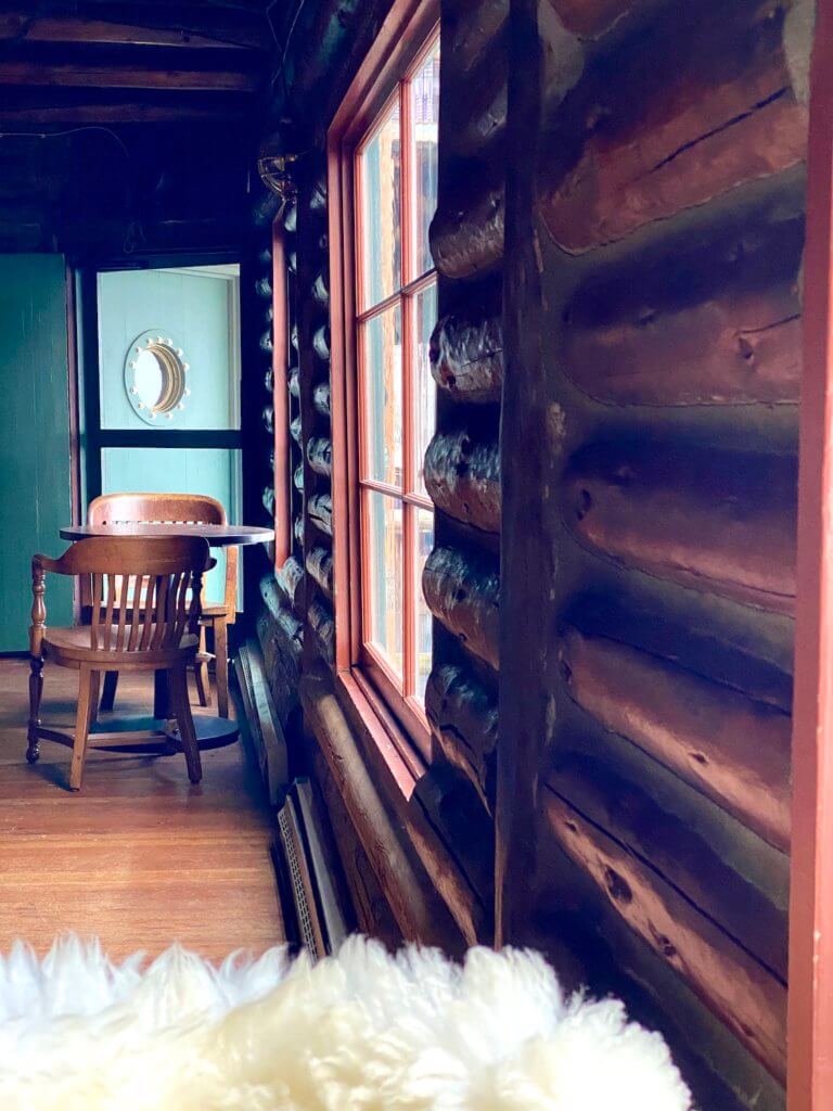 The inside of Captain Whidbey Hotel shows the fluffy white wool blanket draped over a chair while two wooden chairs sit on either side of a wood table, underneath a green door with a porthole. The building is made from logs.