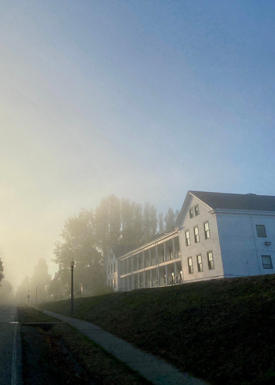 A misty evening comes to life under blue sky with an old style military barracks painted white and double paned windows peeks through the mist at Fort Worden State Park.