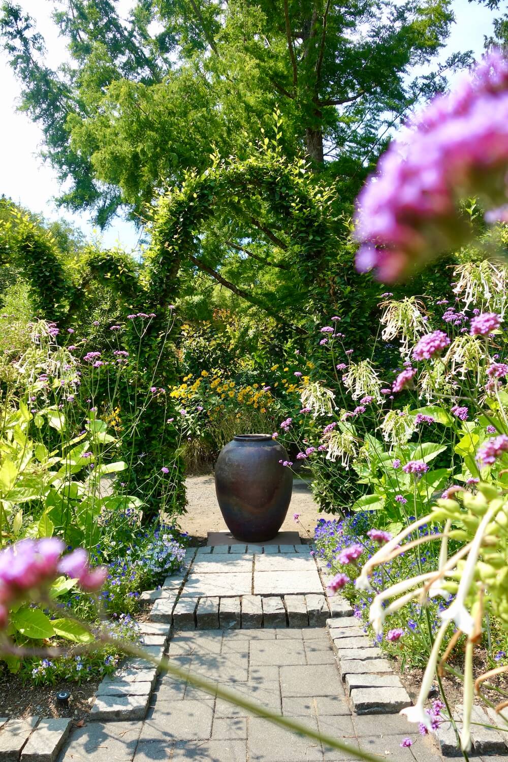 The formal garden at Heronswood near Kingston in Washington State shows elegant green arbor with purple flowers and a large copper urn centrally located amongst a stone pathway. 