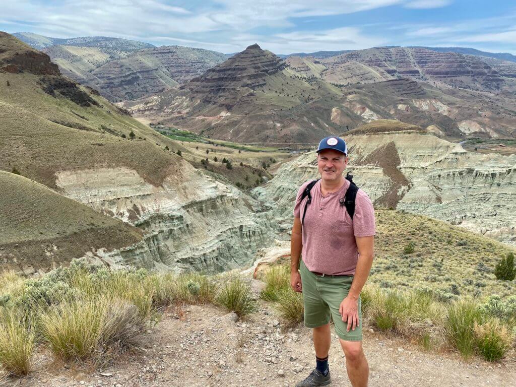 Matthew Kessi poses for a photo at the overlook to the Blue Basin in John Day Fossil Beds National Monument. The canyons are a variety of blue and green colors and continue up to grass covered slopes leading to pointed peaks. There are endless canyons in the background that flow to partly cloudy skies. Matthew is wearing a red shirt and green shorts and a backpack and blue hat. He looks sweaty, as if he is hiking.