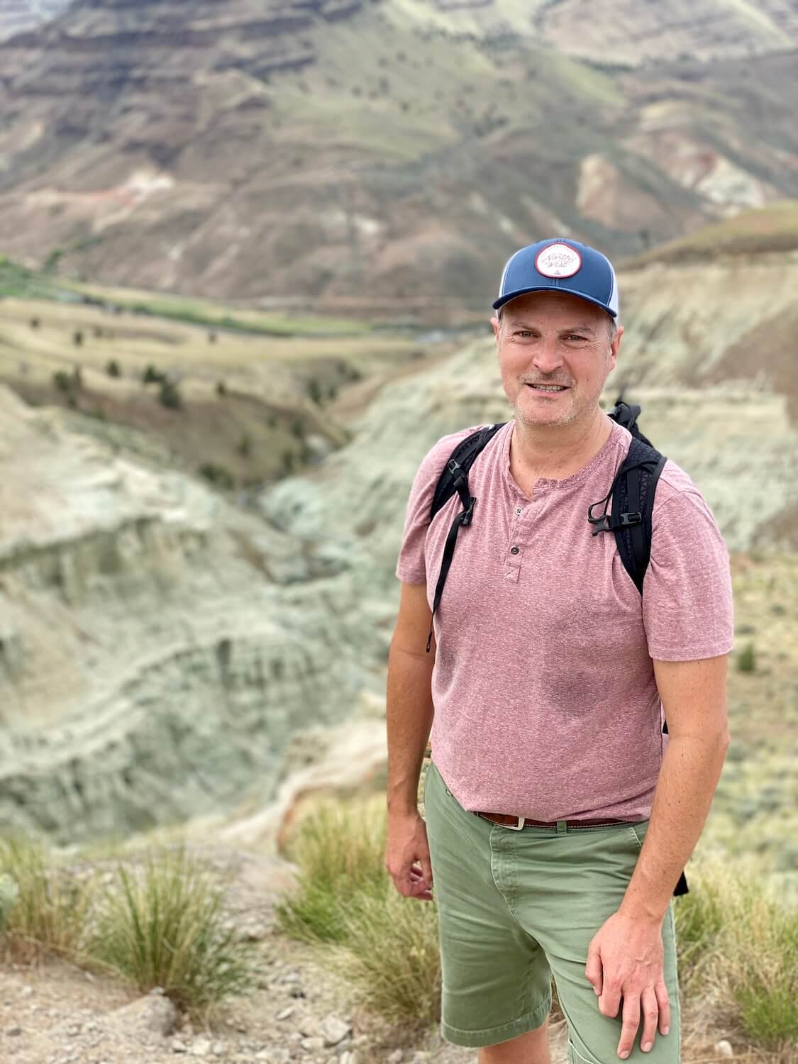 Matthew Kessi poses for a selfie on the Blue Ridge Overlook Trail.  The blue formations of rock are out of focus in the background as he stands there with a red shirt and green shorts.  