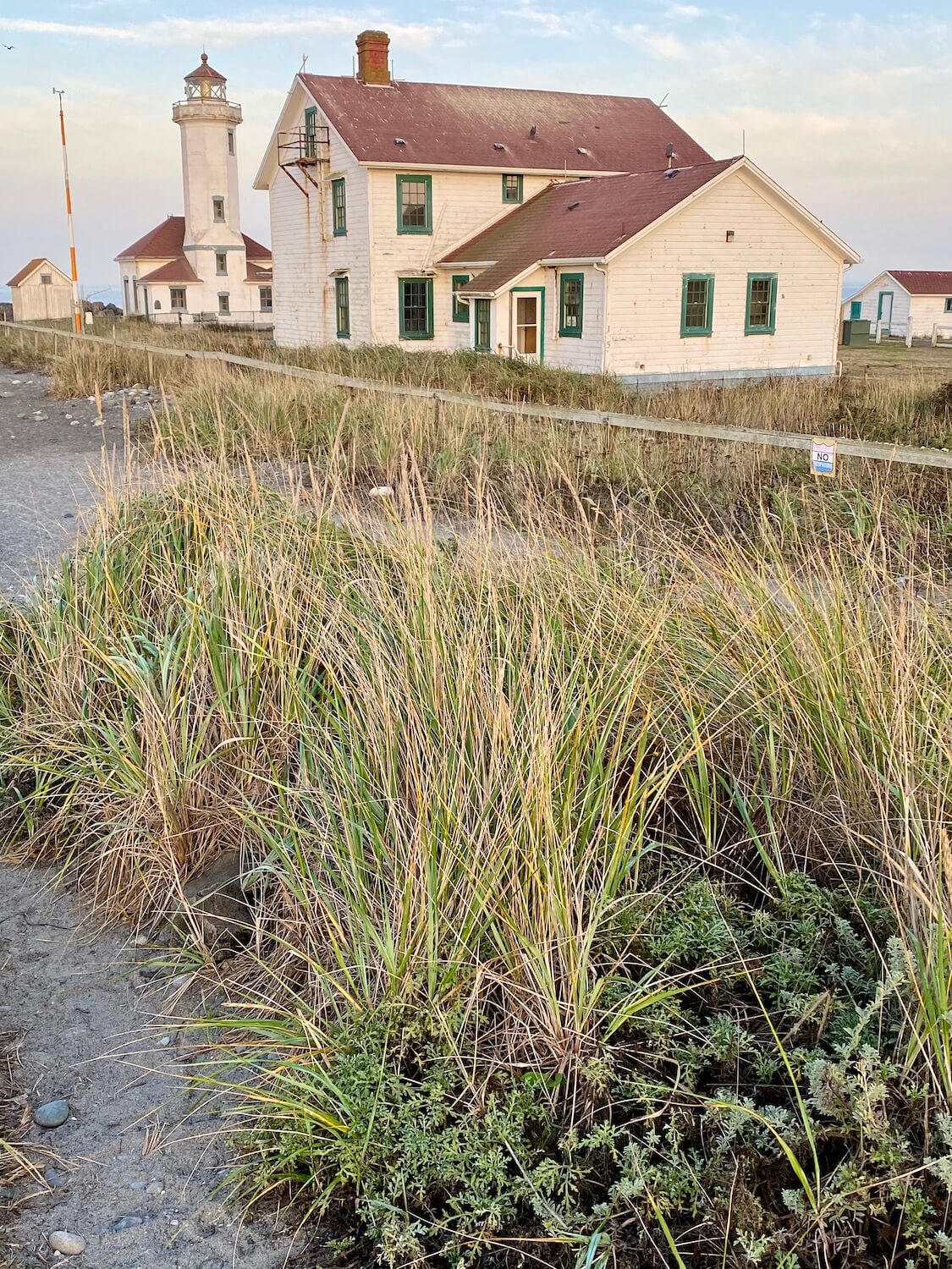 A lighthouse sits in the distance with a red tiled roof and white boarded walls with green trimmed windows. In the near ground is seagrass amongst gray fine sand.