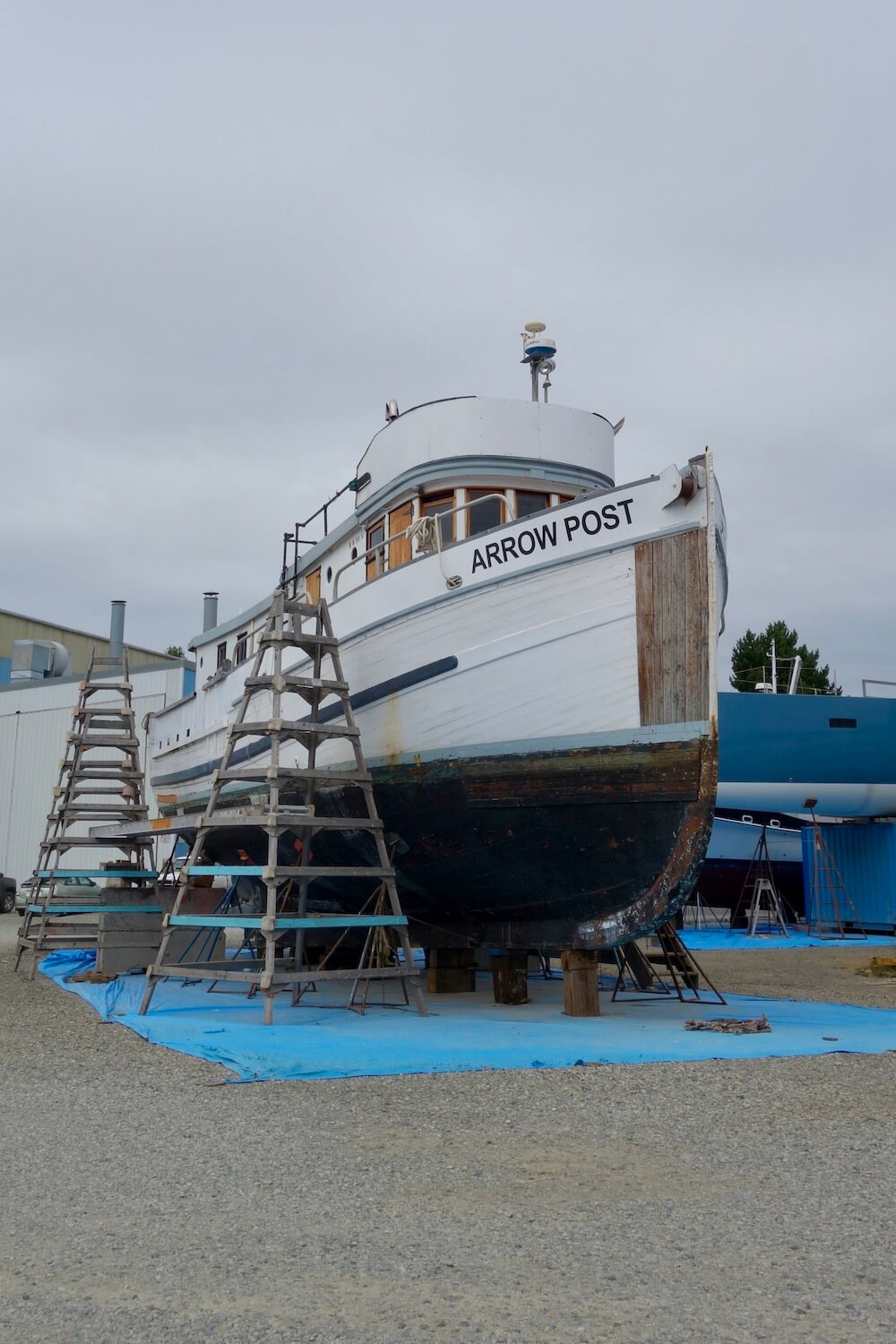 A boat sits, propped up on a dry dock in Port Townsend, Washington. The hull has barnacles and is painted a dull red while above the line the paint is fresh white. The boat's name is Arrow Post and there are several ladders on either side.
