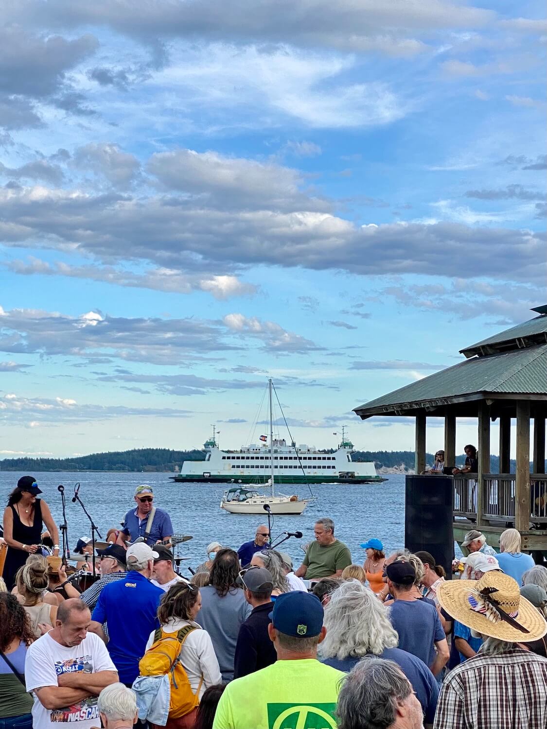 A crowded festival of people dancing at a concert celebrate under partly cloudy skies as a sailboat and ferry glide by in the Salish Sea in the background.