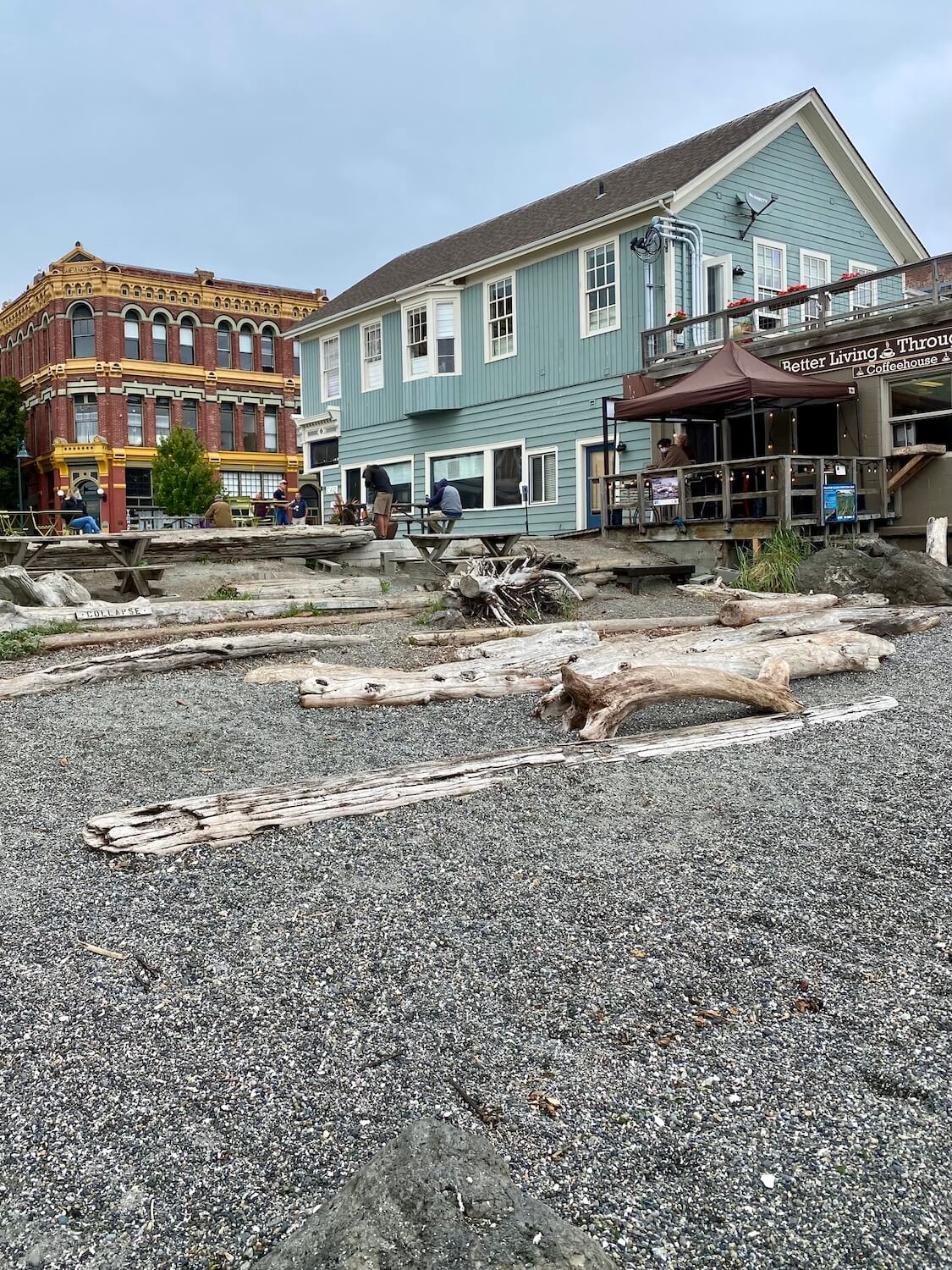 A pebble beach leads up to driftwood that make convenient benches for patrons getting coffee at Better Living through Coffee, a shop right on the beach. In the background you can see an ornate red brick building with bright yellow trim under gray cloudy skies.