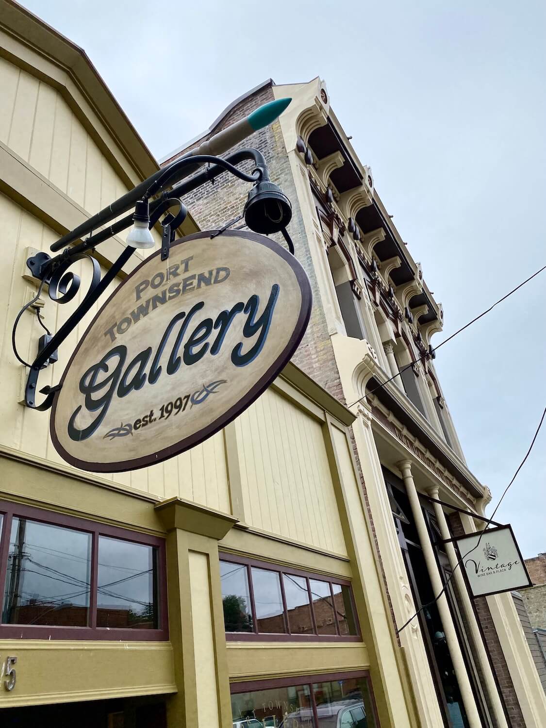 An ornate Gallery sign is posted on a street with yellow and mustard green trim underneath an elegant victorian themed building. Art is a fun thing to do in Port Townsend, Washington