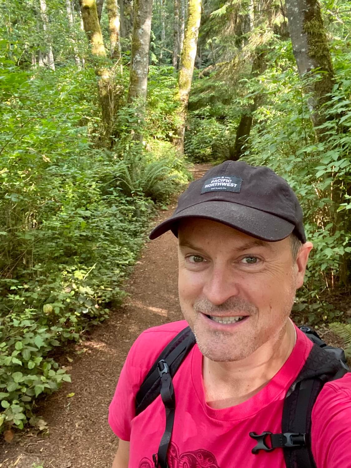Matthew Kessi takes a selfie while hiking in a thickly wooded forest. He's smiling and wearing a bright red shirt and black cap.