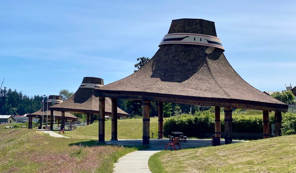 The Swinomish Hat Pavillions are made up of three giant replicas of the traditional cedar hats worn by the Coast Salish People. Around the tops of each structure is a ring with the shape of cedar canoes painted in black outlined by red lines. The sky is blue and the grass is green.
