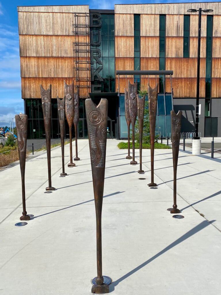 The outside of the Burke Museum has siding made from cedar and large metal letters that spell out Burke. In the foreground is a sculpture made up of 11 bronze paddles used by people of the Columbia River.