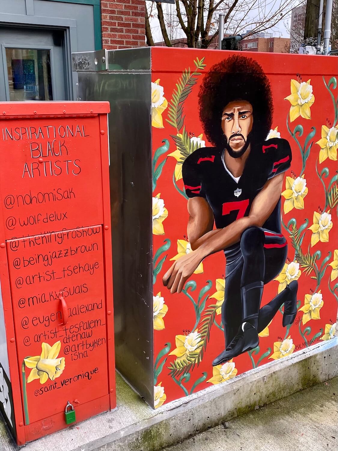 This mural on an electrical panel on Capitol Hill in Seattle depicts Colin Kaepernick kneeling in his football jersey amongst red flowers with yellow daffodils. There is another panel painted red with a list of Inspirational Black Artists.