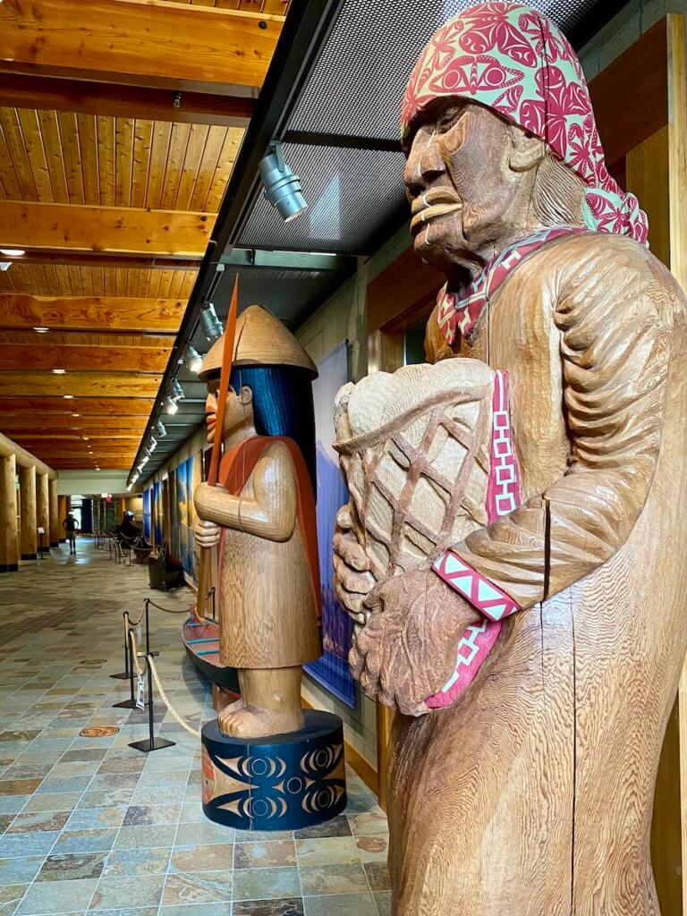 Large wooden carved statures of figures welcome guests to the Hibulb Cultural Center, which is on the Tulalip Indian Reservation. The figure in the foreground is holding a basket of clams and wearing a fabric hood with red and green coloring.