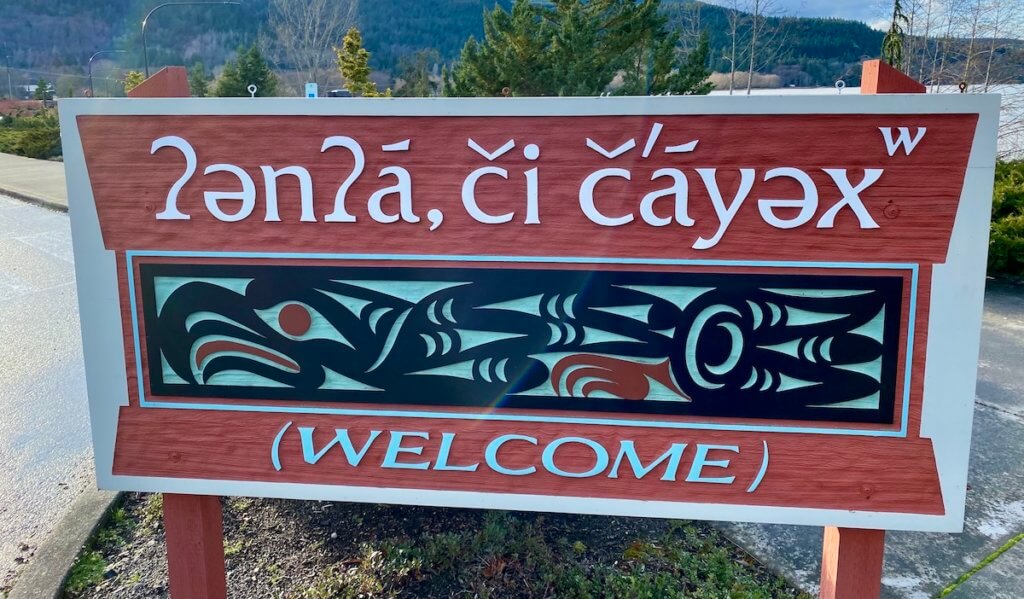 A sign welcoming people to the Jamestown S'Klallam Tribal center near Sequim, Washington. The sign is a brilliant red color with Lushootseed letters in white and English letter in blue. In the middle is Coast Salish art with black, green and red coloring.