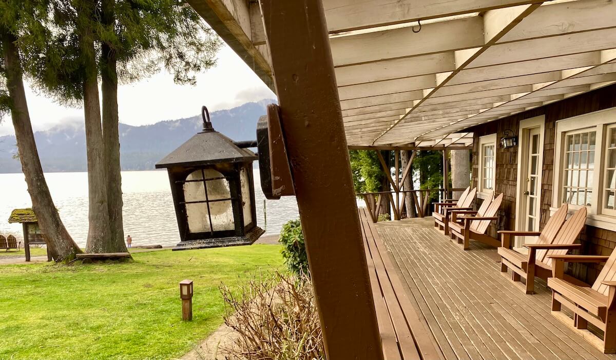 Lake Quinault Lodge is a classic Pacific Northwest hotel and a unique place to stay in Washington State. This is the view of the lake from the boat house, with wood decking and chairs along a wall with paned windows. The lawn is green with grass and an old fashioned light hangs on a post.