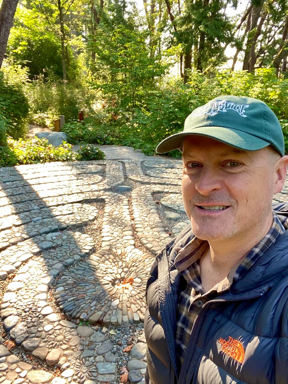 Matthew Kessi smiles for a selfie in front of a Labyrinth on Bainbridge Island. The Labyrinth is made of stones collected from the island and this is under the canopy of a fir forest.