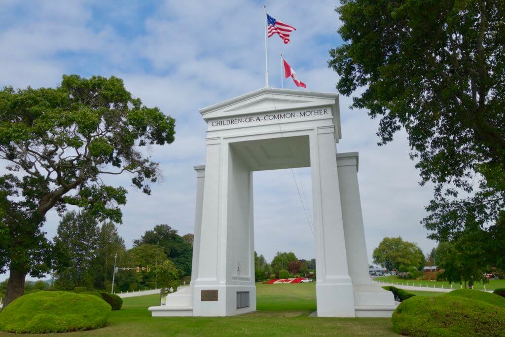 The giant Peace Arch at the United States Canada border is a milestone along the Seattle to Vancouver Drive. The giant white marble columns are located on a grassy green lawn with trees and gardens and both the US and Canadian flags fly proudly overhead.