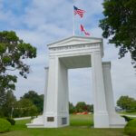 The giant Peace Arch at the United States Canada border is a milestone along the Seattle to Vancouver Drive. The giant white marble columns are located on a grassy green lawn with trees and gardens and both the US and Canadian flags fly proudly overhead.