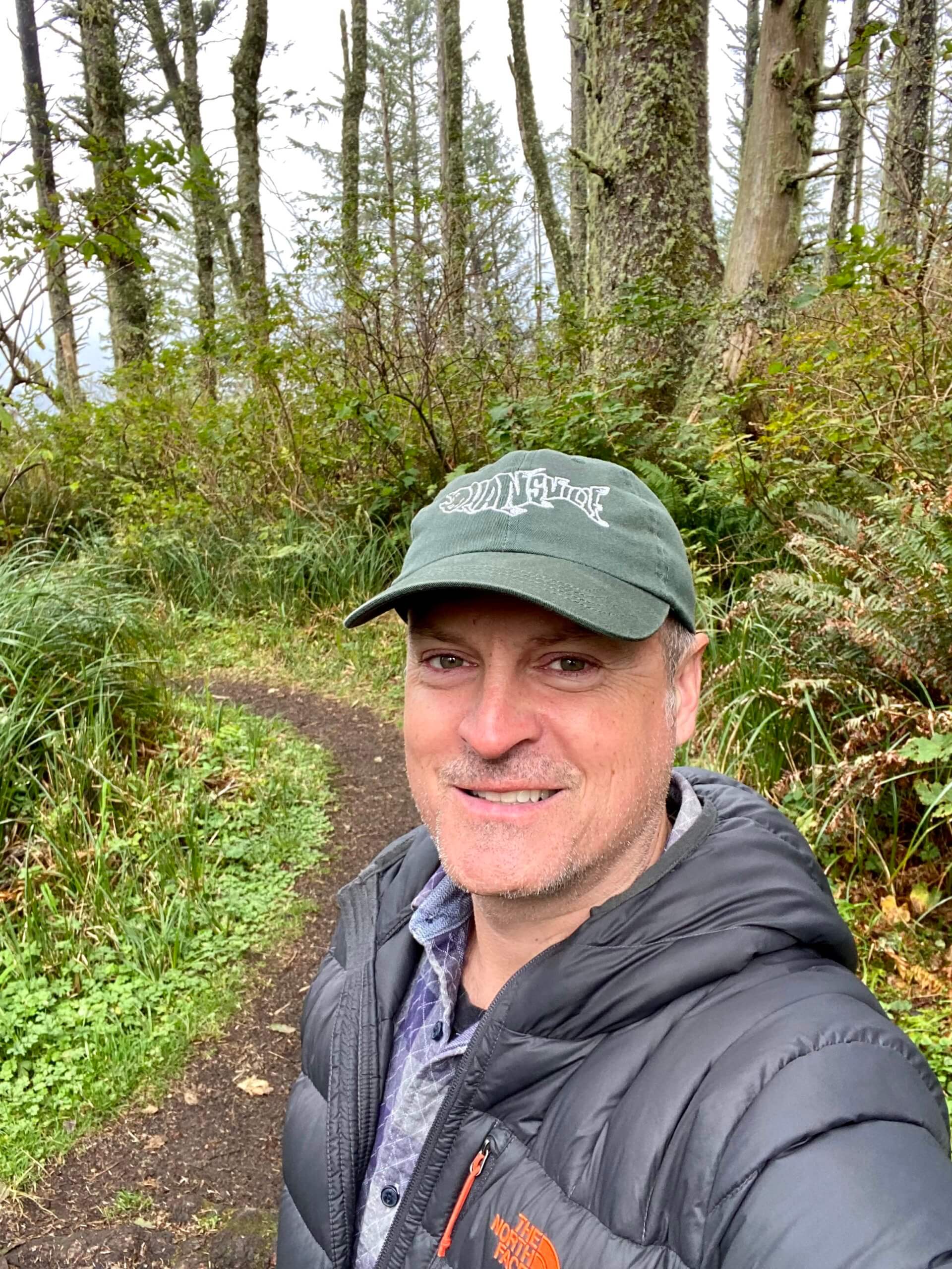 Matthew Kessi poses for a selfie in a forest near Yachats Oregon. He's smiling while wearing a green cap and gray puffy coat while a forest of sitka spruce trees pop up along the hiking trail behind him.