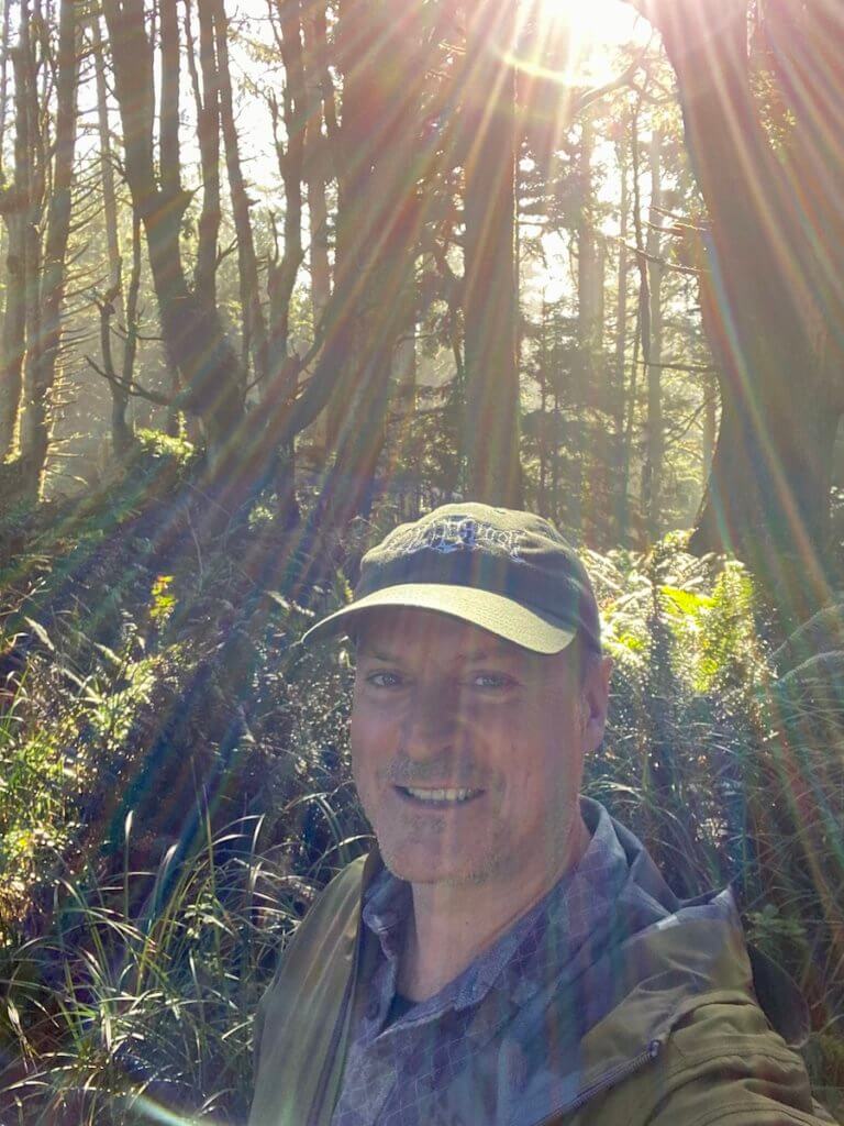 Matthew Kessi poses for a selfie under the rainbow sunbeams filtering through the forest. He's smiling and wearing a green cap and jacket.