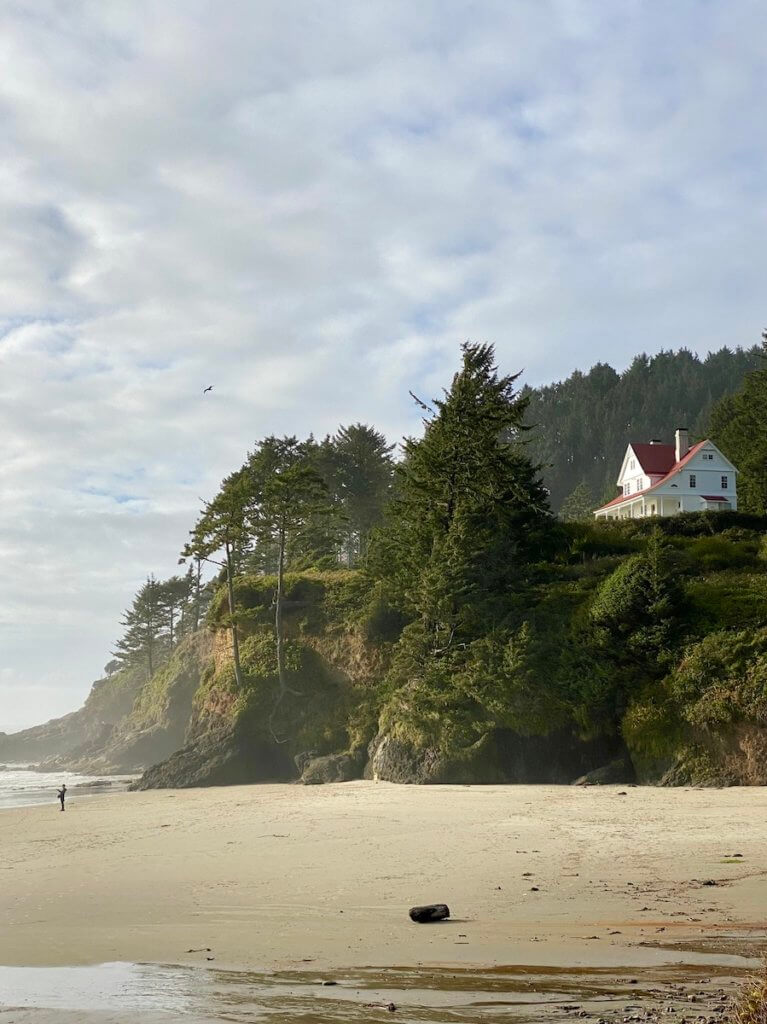 The beach at the base of Heceta Head Lighthouse. On the bluff the bed and breakfast is visible with white outside and red roof and trim. There are rocks on the beach that lead to smooth sand and the waves in the distance.