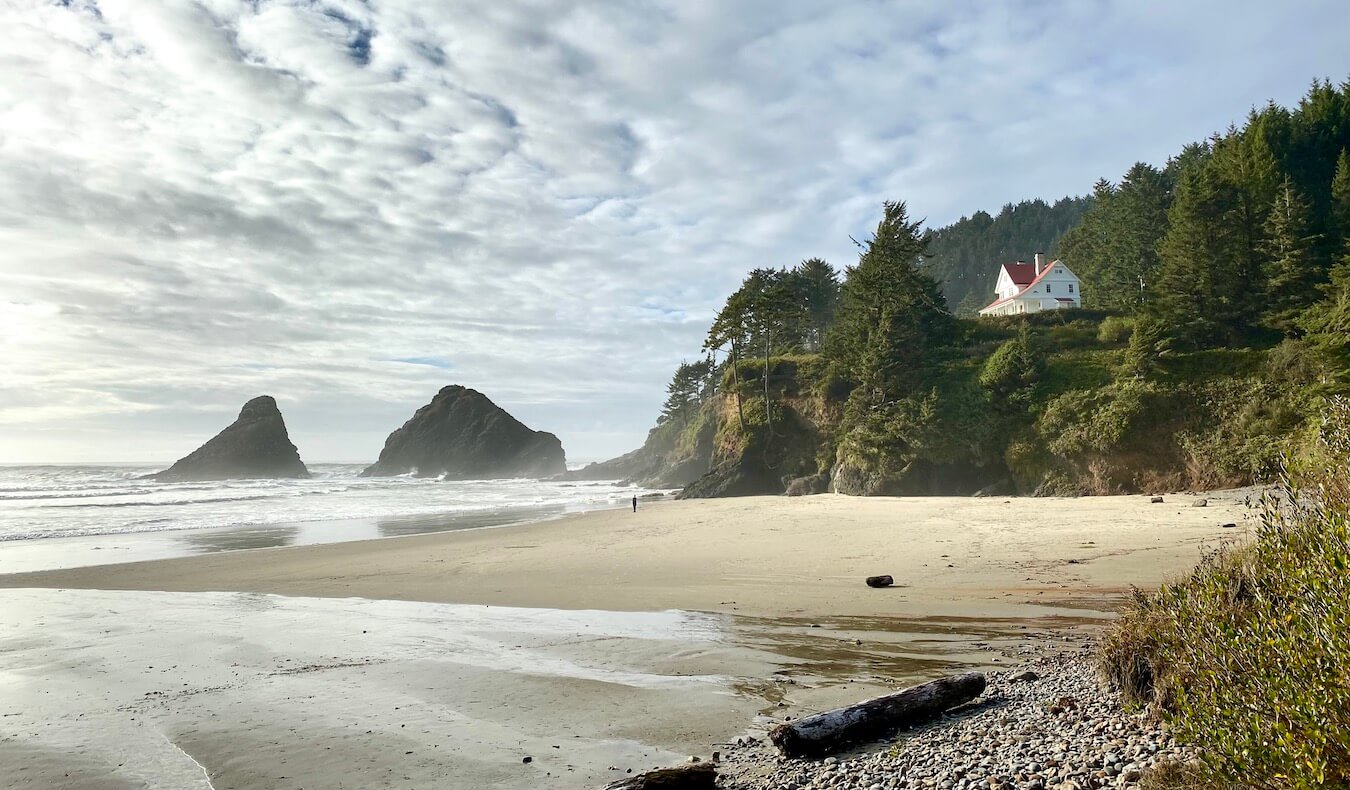 The beach at the base of Heceta Head Lighthouse. On the bluff the bed and breakfast is visible with white outside and red roof and trim. There are rocks on the beach that lead to smooth sand and the waves in the distance.