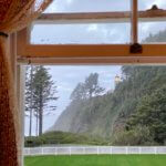 View from a bedroom window looking at the shining light of the Heceta Lighthouse. The yard in the foreground is full of green grass and surrounded by a white picket fence.