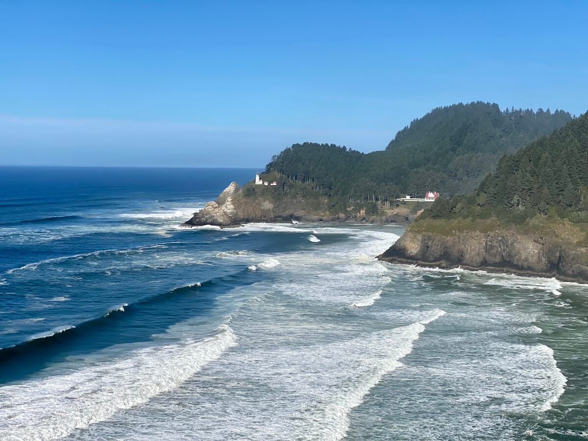 Oregon Coast scene featuring Heceta Head Lighthouse and the nearby Bed and Breakfast with layers of surf floating in and pounding on the rocks. The mountains rise up, covered in evergreen trees.