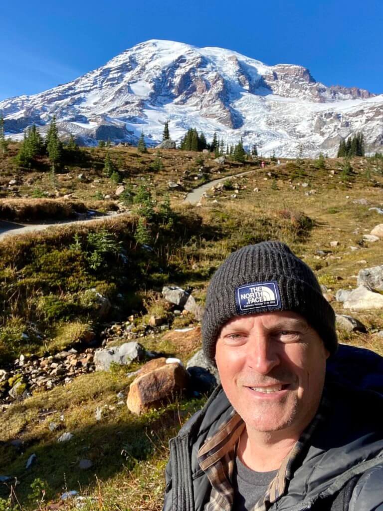 Matthew Kessi poses for a selfie while hiking on a trail on Mt. Rainier. The mountain is covered in snow and pops against the blue sky.