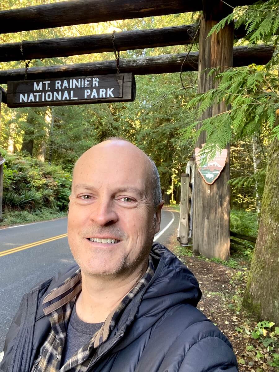 Matthew Kessi poses for a selfie in front of the entrance sign to Mt. Rainier National Park. He is along the paved road with yellow lines framed in by cedar fir trees.