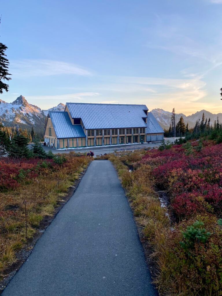 The Visitor Center at Paradise on Mt. Rainier is shown at the end of a path surrounded by low brush changing colors for fall. The sky is blue.