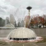 This shot shows Seattle in November with the trees around the Seattle Center changing leaves and dropping them with the Space Needle hovering above in cloudy skies. In the foreground is the International Fountain spraying water streams in every direction, with the water bubbling down to the drain, which is red brick color.