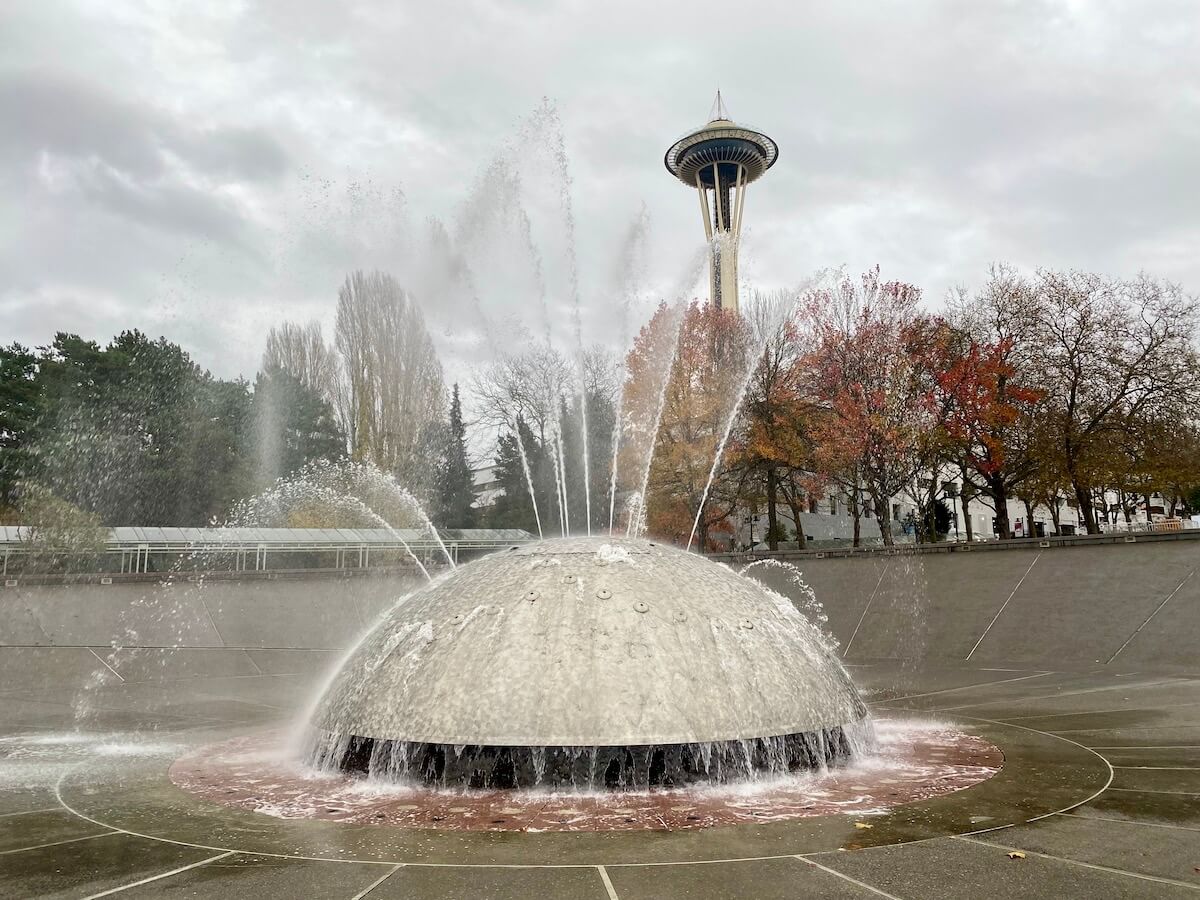 This shot shows Seattle in November with the trees around the Seattle Center changing leaves and dropping them with the Space Needle hovering above in cloudy skies. In the foreground is the International Fountain spraying water streams in every direction, with the water bubbling down to the drain, which is red brick color.