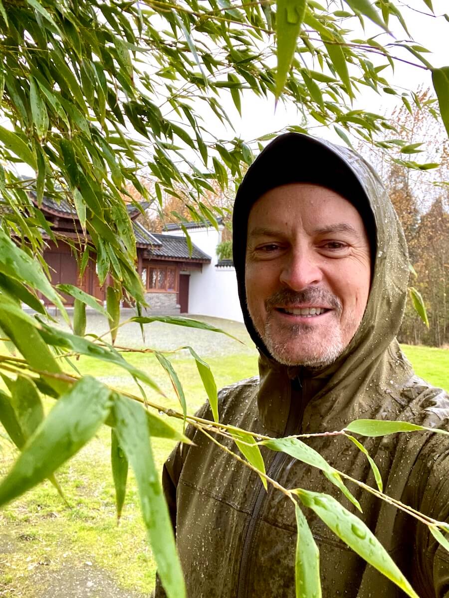 Matthew Kessi smiles for a selfie, covered in water from a rainstorm. He's standing under a bamboo plant with a Chinese pagoda in the background.