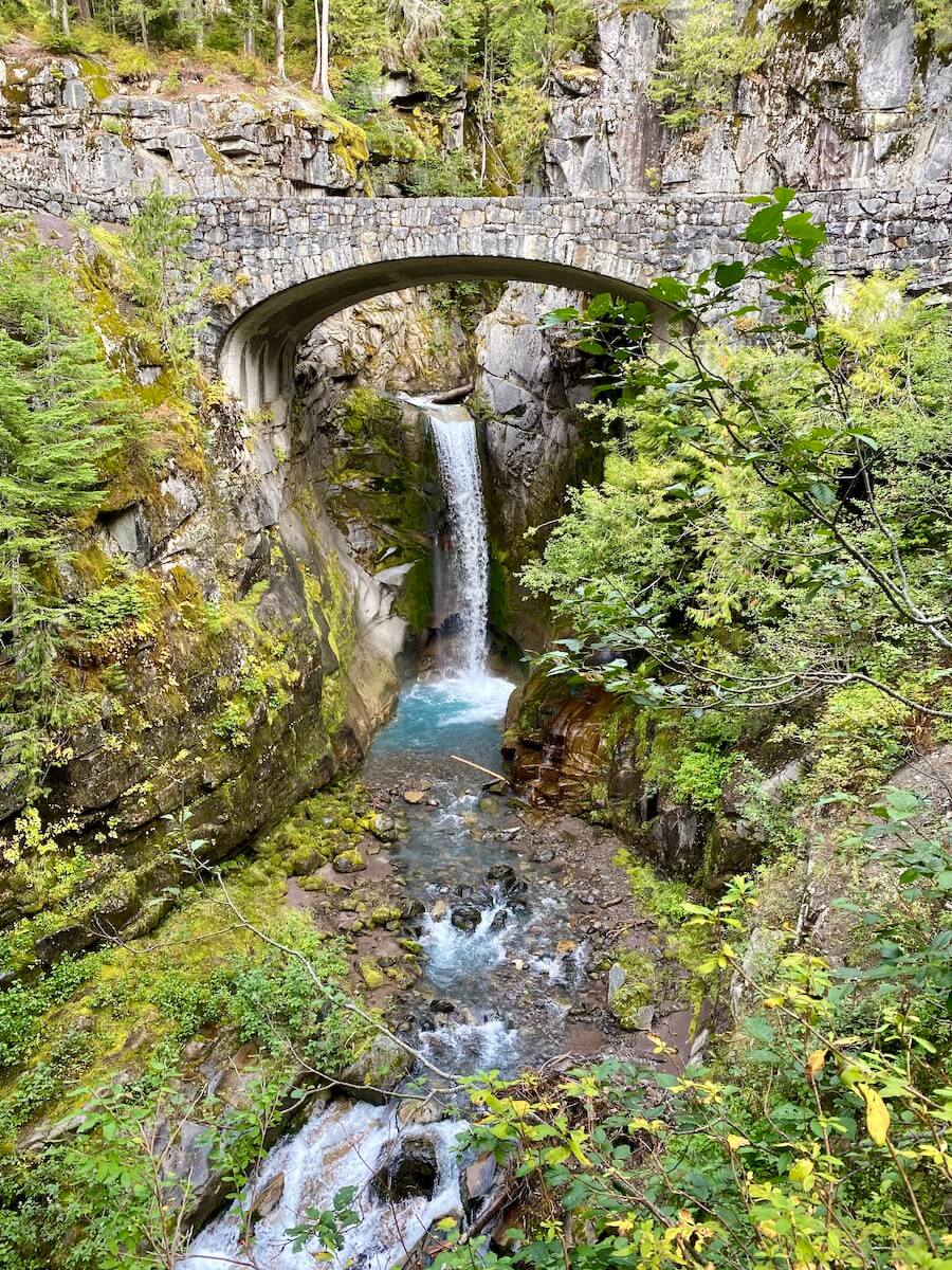 A beautiful waterfall cascades down a mountain side underneath a stone roadway bridge. Various green fir trees and bushes frame in the shot.