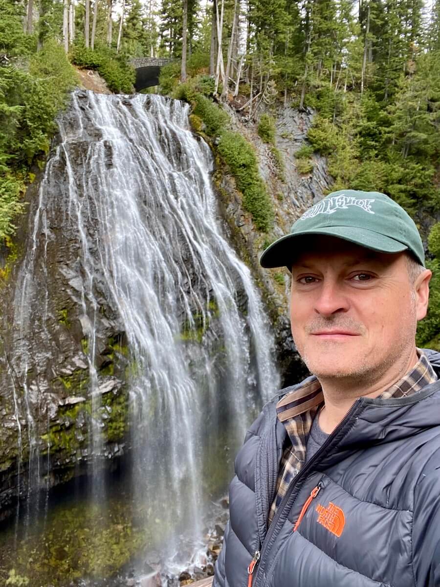 Matthew Kessi stands for a selfie in front of a flowing waterfall that seems to drape down basalt lava cliffs on Mt. Rainier.