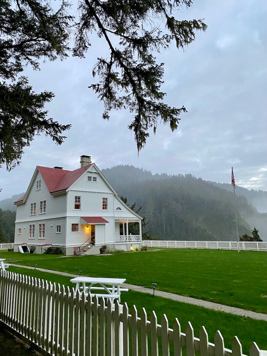 The Heceta Lighthouse Bed and Breakfast is an old wooden building painted white with a red roof and a large green lawn surrounded by a white picket fence. Mist rises up from a forest in the background.