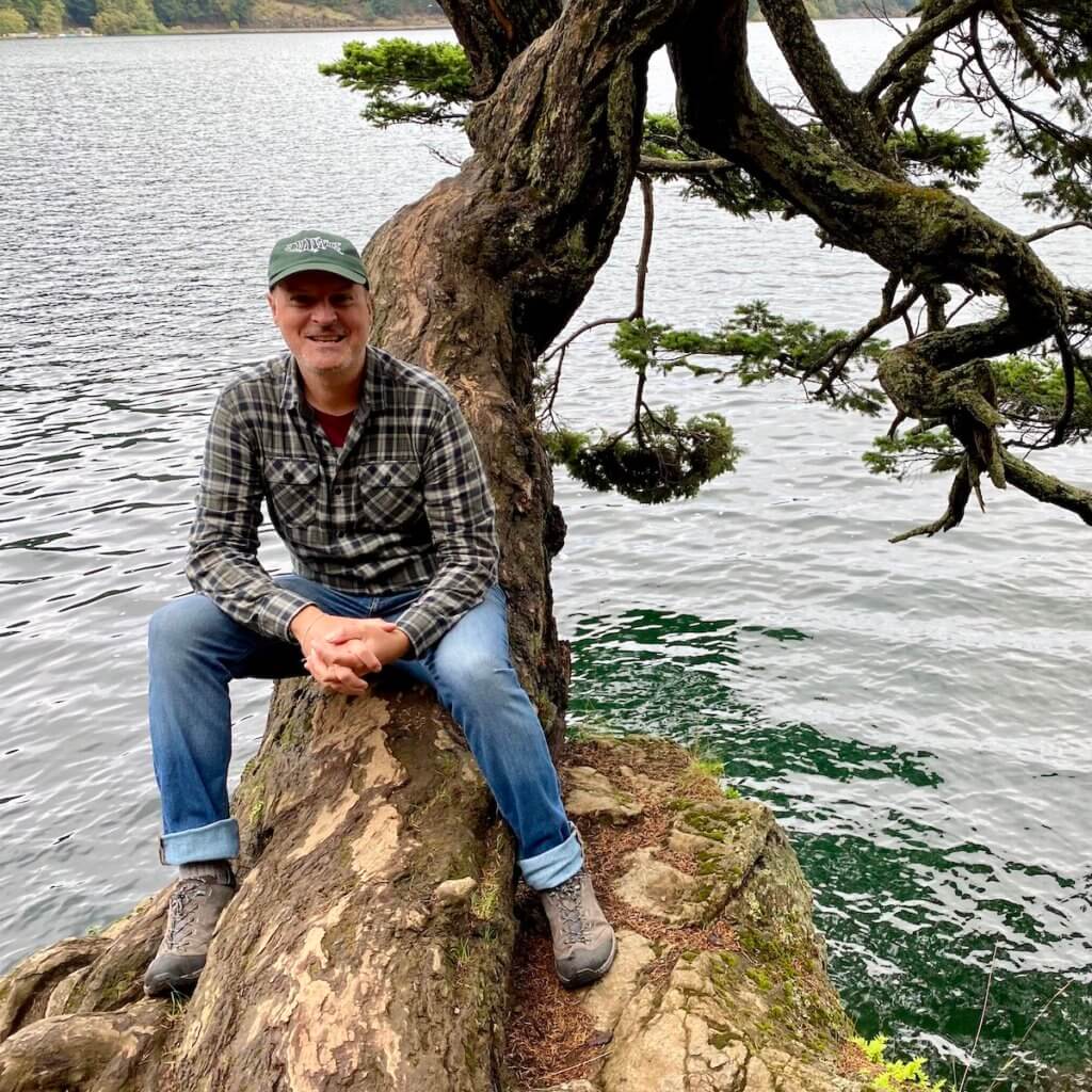 Selfie of Matthew Kessi sitting on a tree stump overlooking a lake with greenish water.