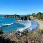 Chetco Point Park is a great thing to do in Brookings Oregon. Waves roll into shore with bright white caps, smothering over an open sandy beach while blue skies float above this scene.