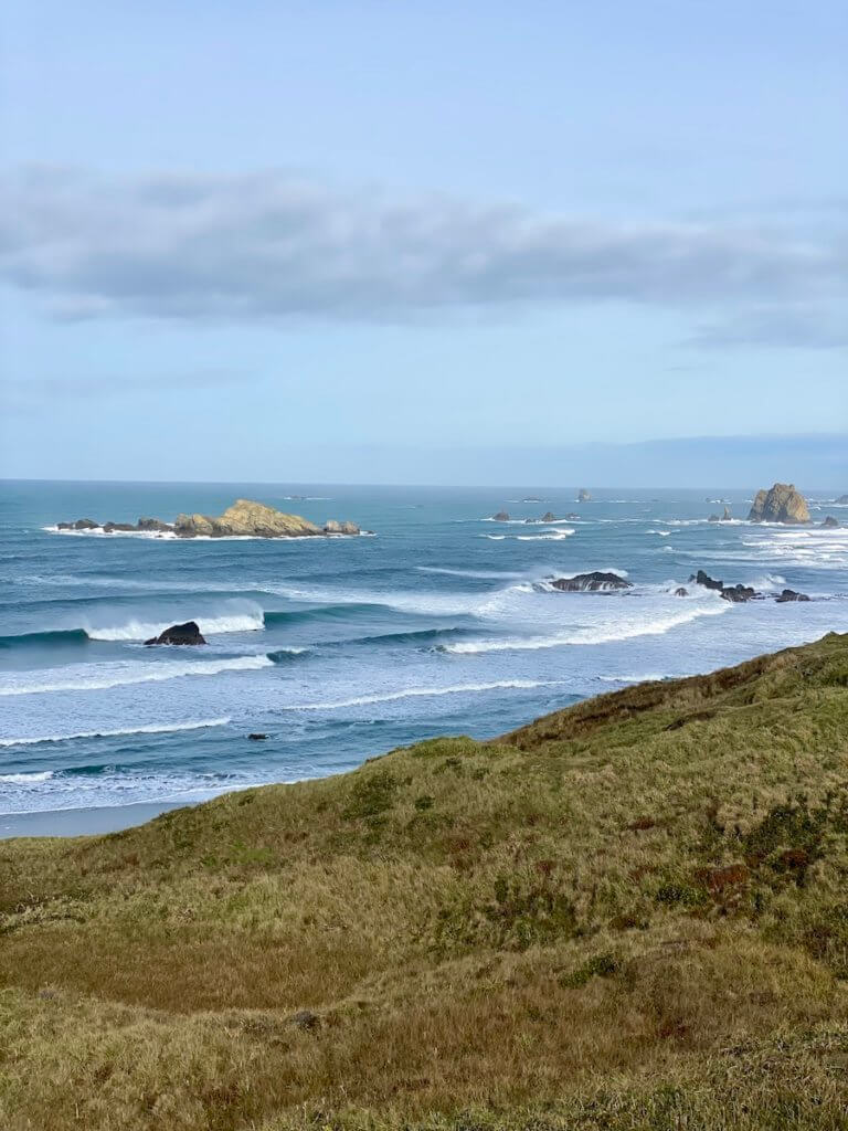 Soulful travel on the Oregon Coast involves sweeping views of the surf like in this picture, where bright white waves crash against rocks along the coastline under blue skies.