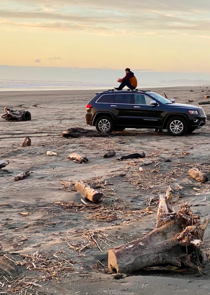 A man sits on top of a Jeep Cherokee vehicle and looks out at the scenic horizon of the Oregon Coast. The sun is setting, providing a myriad of yellow and light blue colors in the sky.