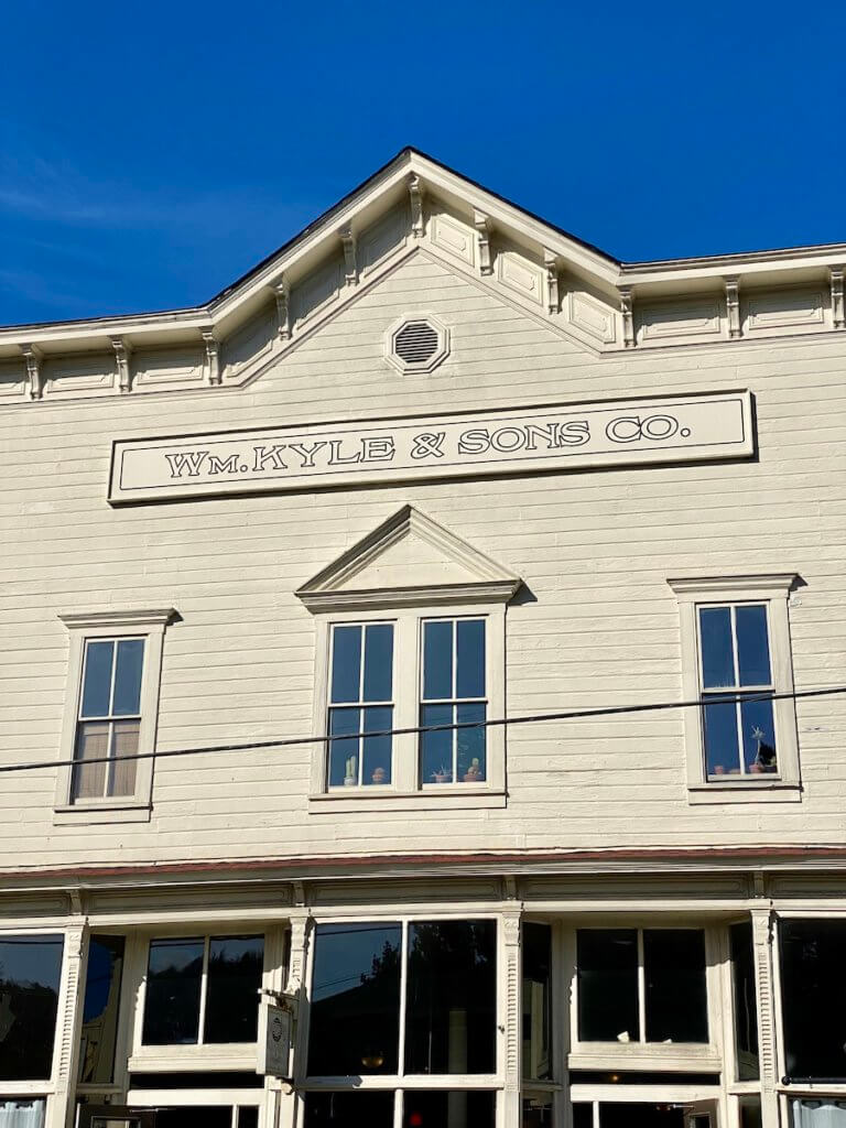 An old pioneer style store front rises up in historic Florence Oregon and offers a glimpse into the past along the Oregon Coast. The sky is bright blue above and the wooden siding is painted a cream color.